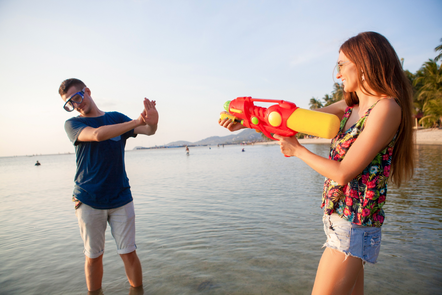 Young woman playfully spraying a man with a water gun at the beach, enjoying island life and outdoor fun.
